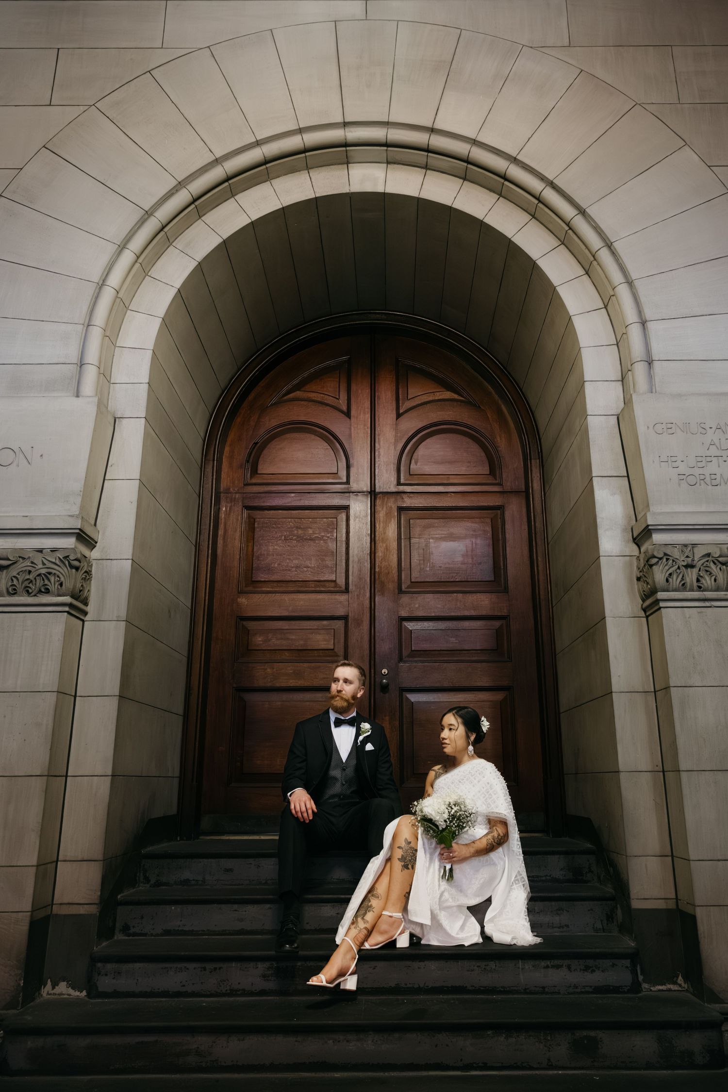 Bride and groom sitting on steps beneath grand arched wooden doors of historic building in elegant wedding attire.
