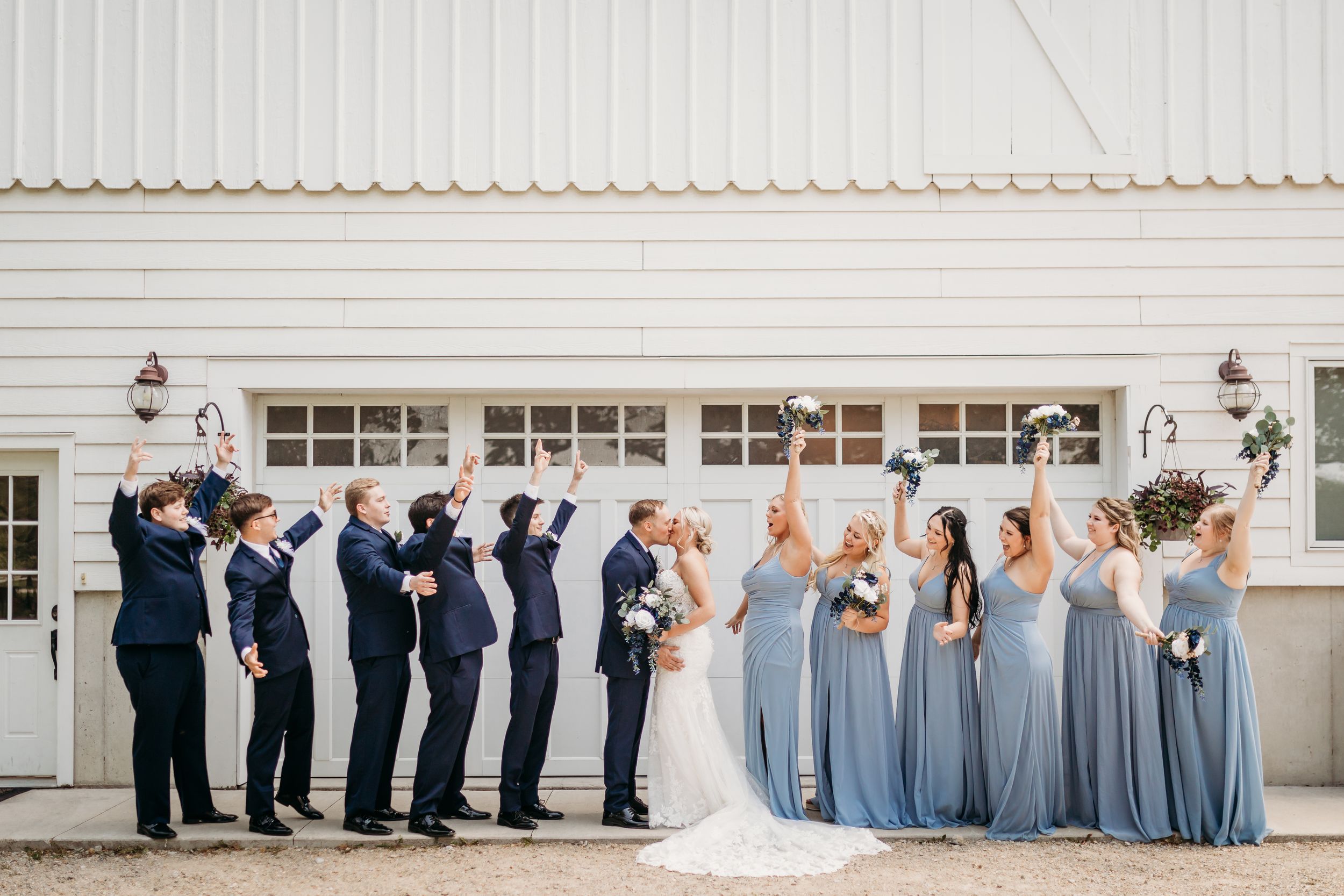 Wedding party in navy suits and blue gowns cheering outside a white barn venue.