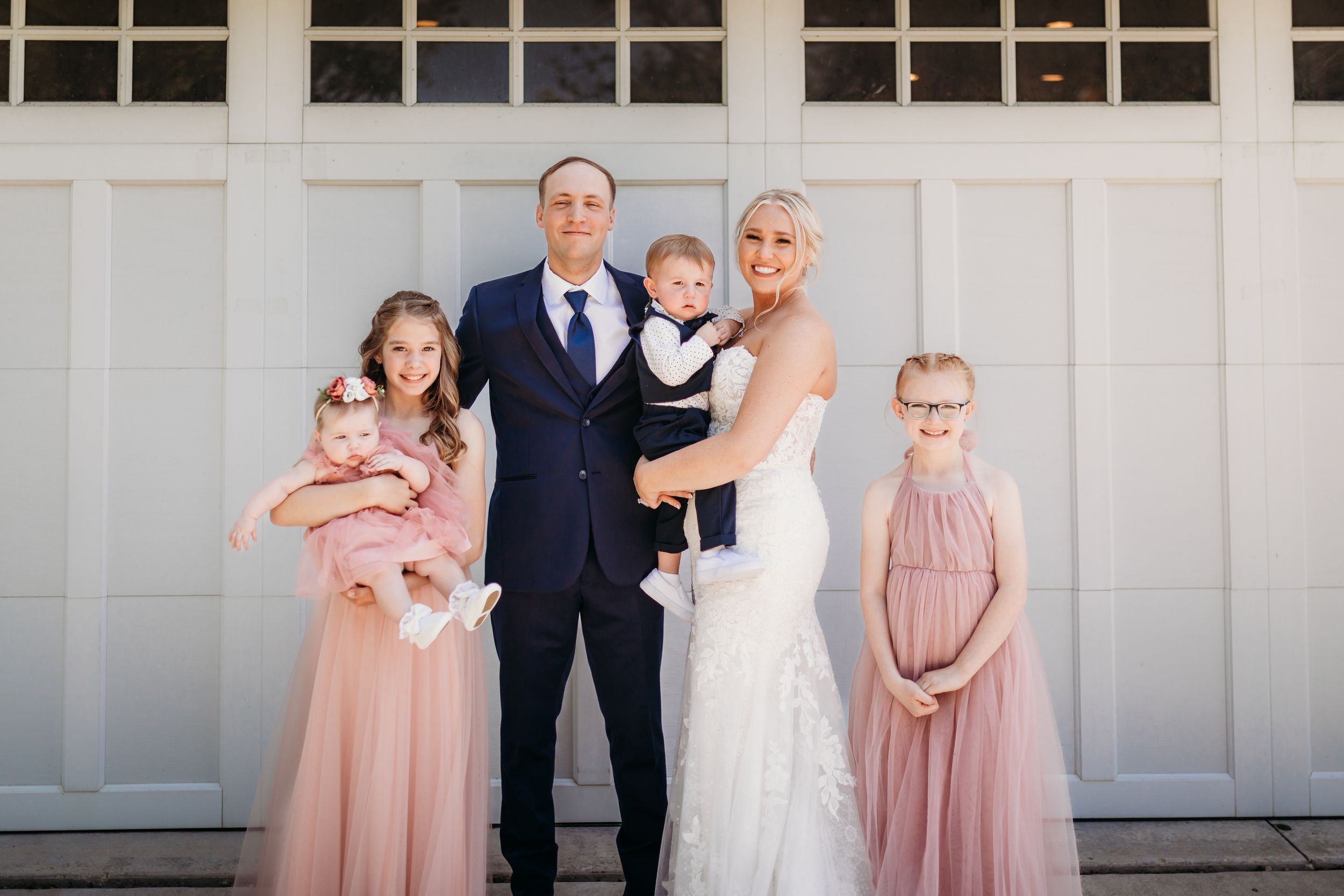 Family portrait at wedding with groom in navy suit, bride in lace gown, and three children in blush pink dresses.
