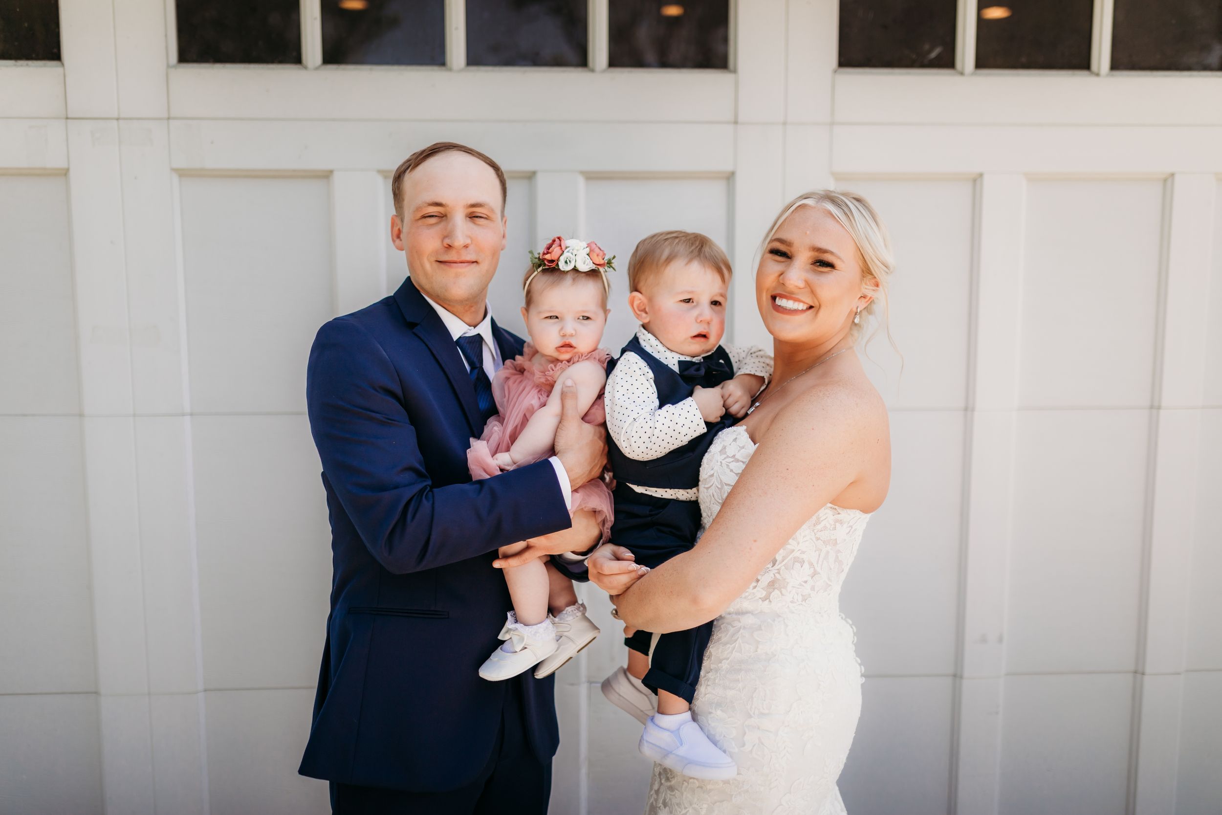 Bride and groom in wedding attire hold two young children in front of a white barn venue.