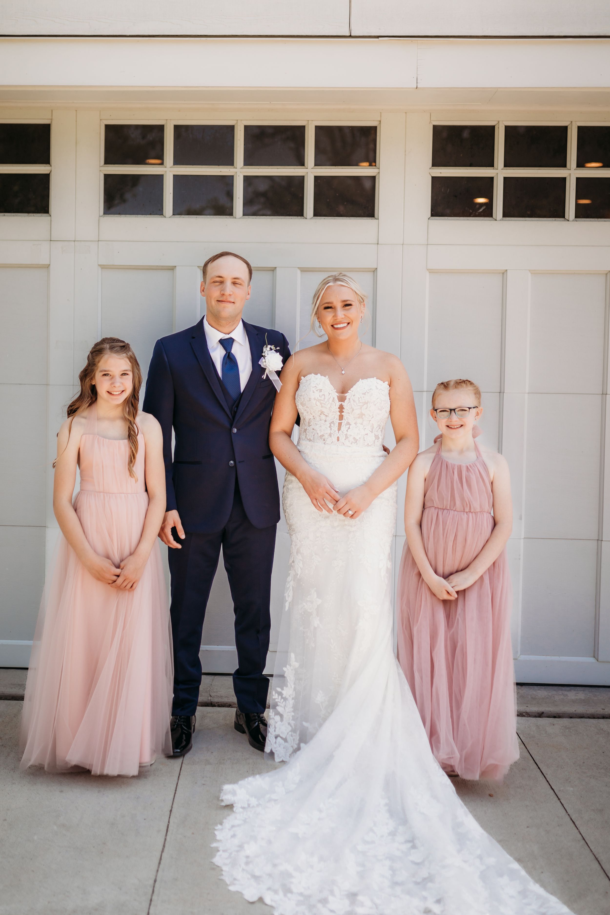 Bride and groom pose with two flower girls in pink dresses before a garage door.