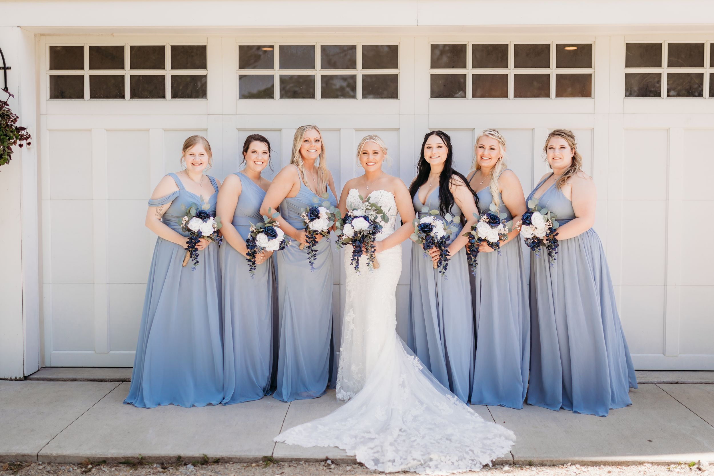 Bride in lace wedding gown poses with six bridesmaids in dusty blue dresses holding floral bouquets.