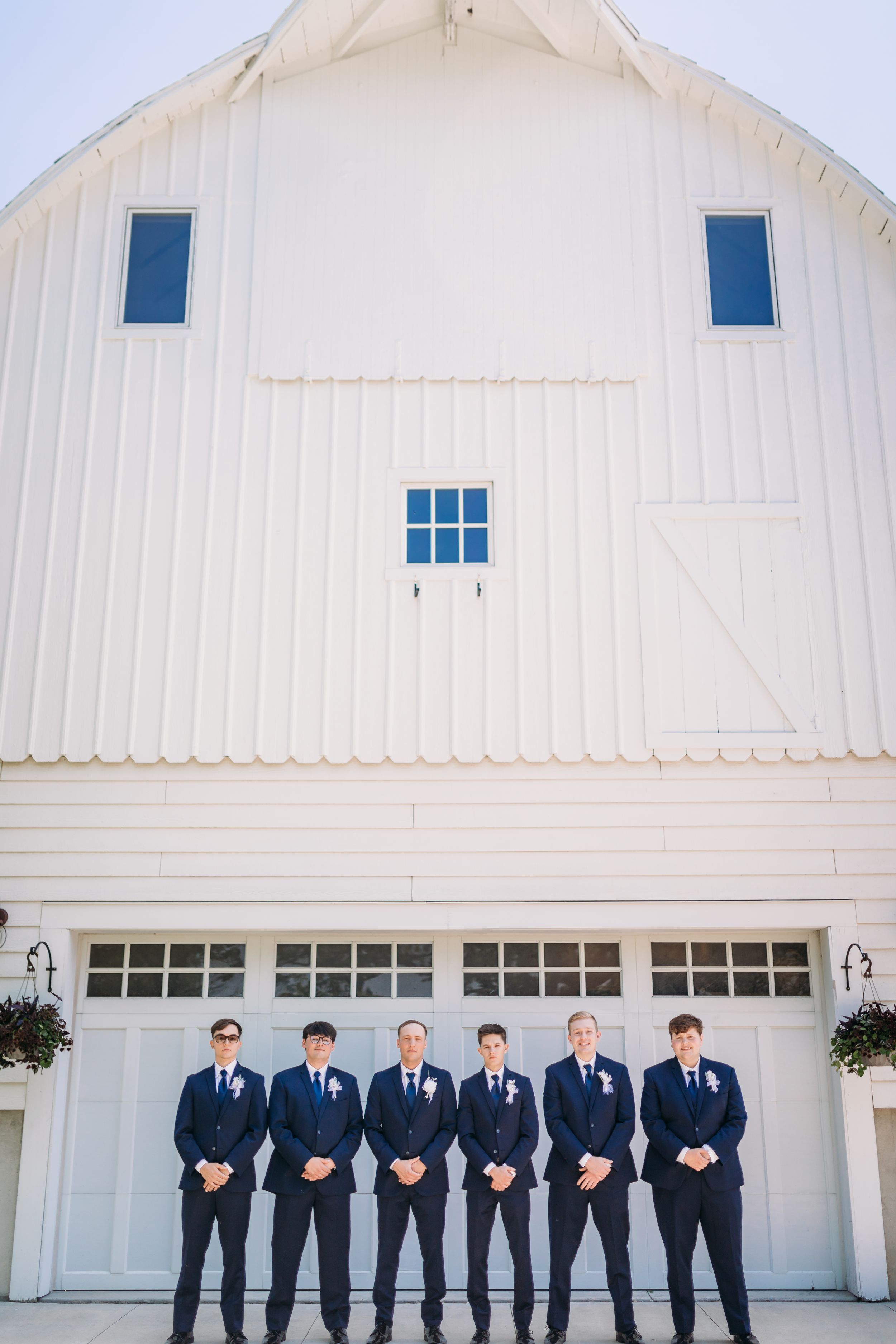 Six groomsmen in navy suits with boutonnieres stand in front of a white barn wedding venue.