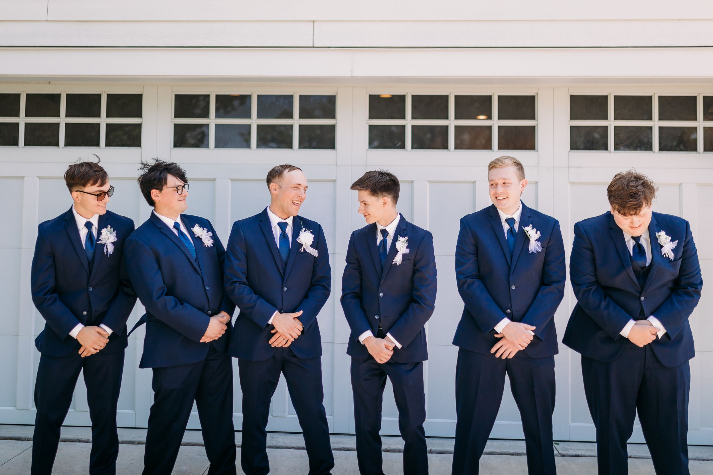 Six groomsmen in navy blue suits with floral boutonnieres posing in front of a white garage door at a wedding.