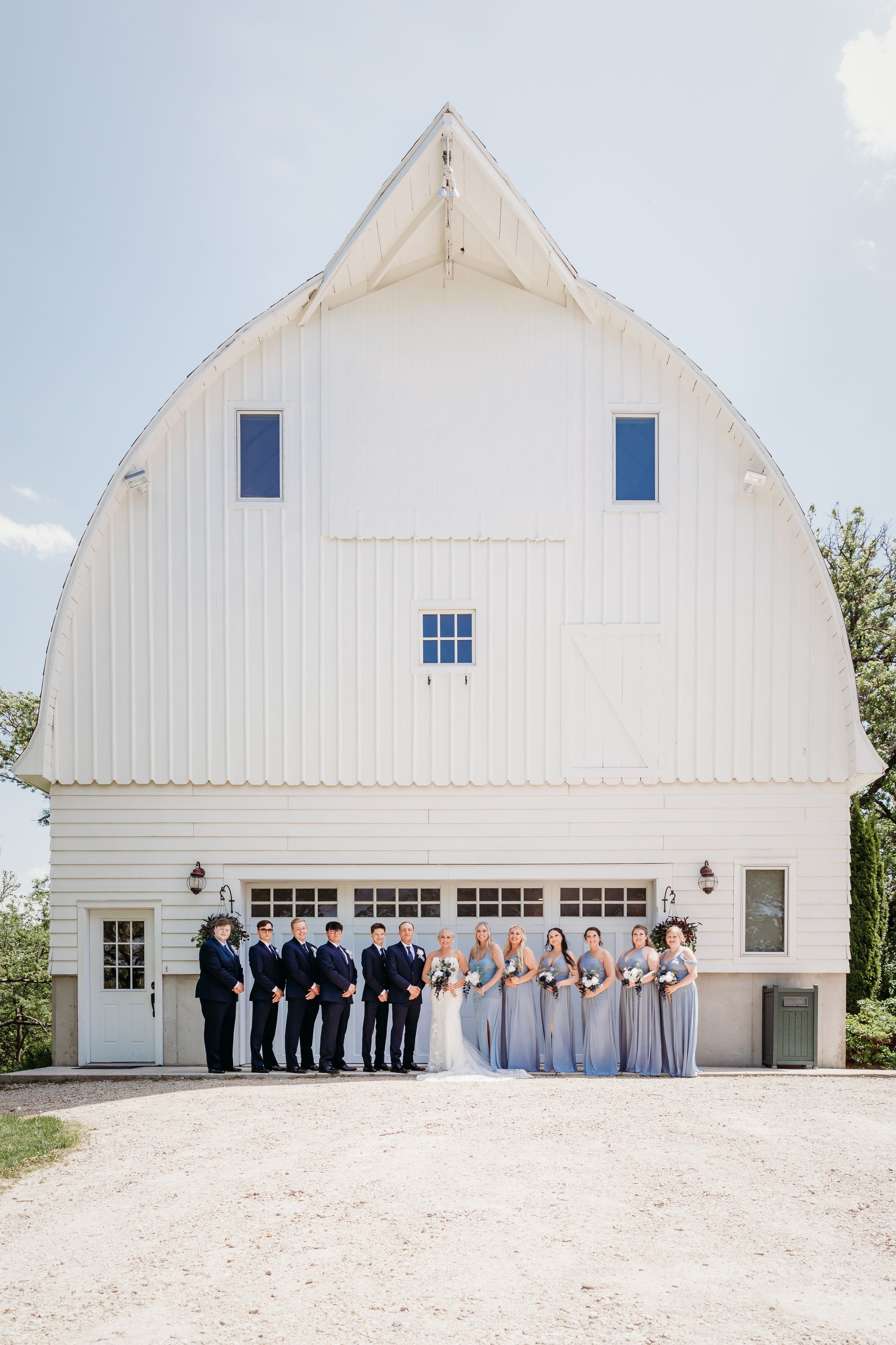 Wedding party posing in front of a white barn venue on a sunny day, bridesmaids in dusty blue gowns.