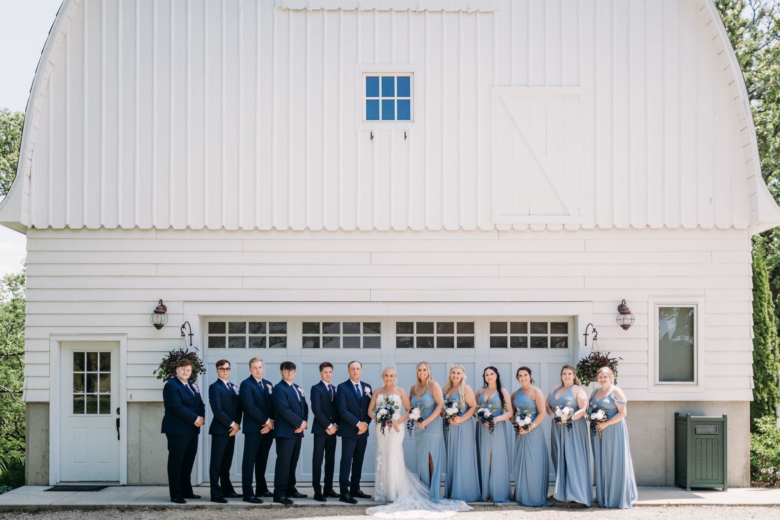 Wedding party poses in dusty blue and navy attire before a white barn venue.
