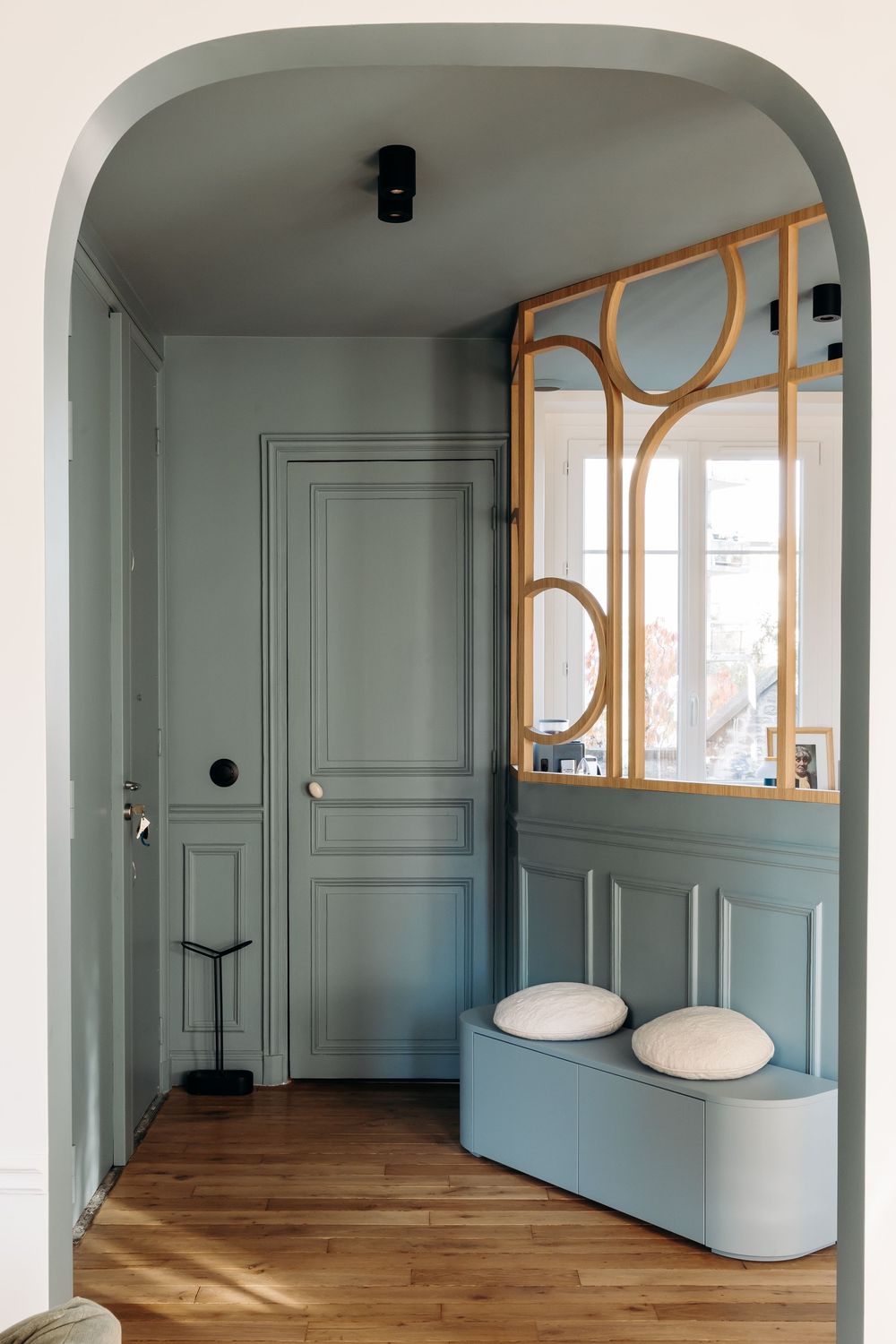 Elegant entryway with sage green paneled walls, arched doorway, rattan mirror partition, and a light blue storage bench on hardwood floors.