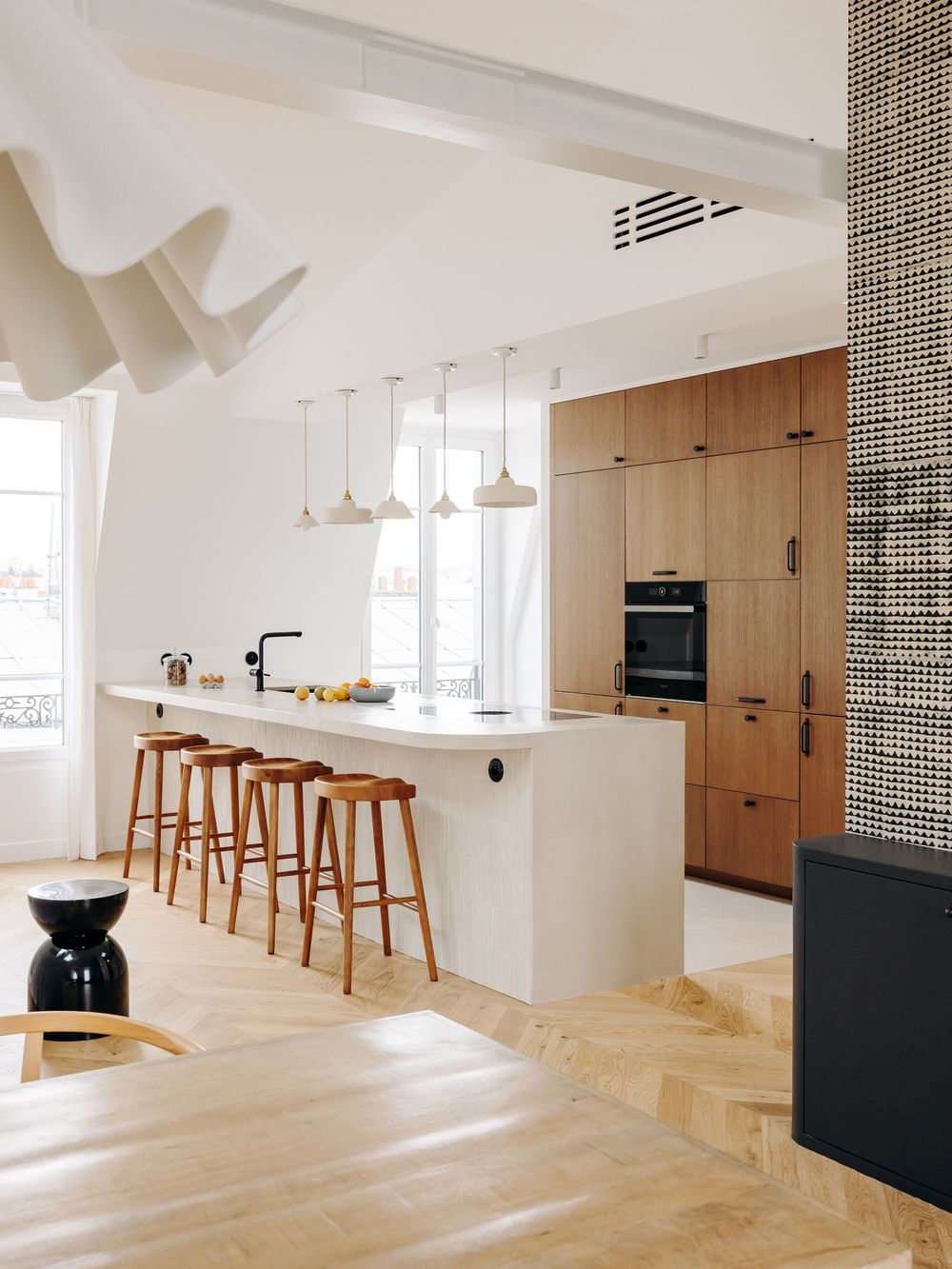 Modern Scandinavian kitchen with white island, wood bar stools, oak cabinetry, herringbone floor, and pendant lights.