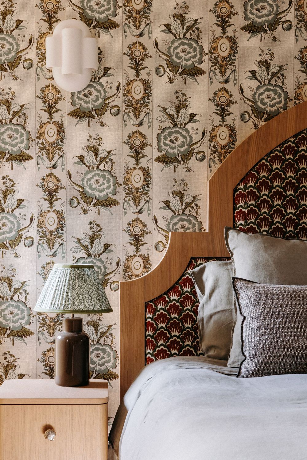 Elegant bedroom with floral wallpaper, arched wooden headboard, patterned upholstery, neutral pillows, and a green lamp on nightstand.