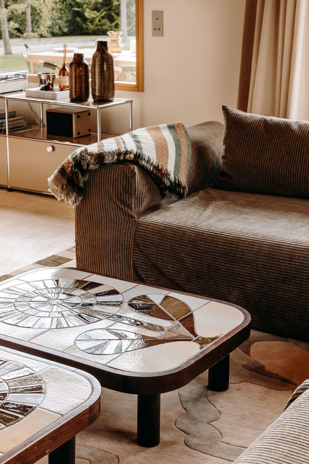 Cozy living room with corduroy sofa, striped throw, mosaic coffee tables, and warm neutral decor near a sunlit window.
