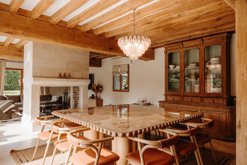 Rustic dining room with wooden beam ceiling, marble table, wicker chairs, crystal chandelier, and antique hutch cabinet.