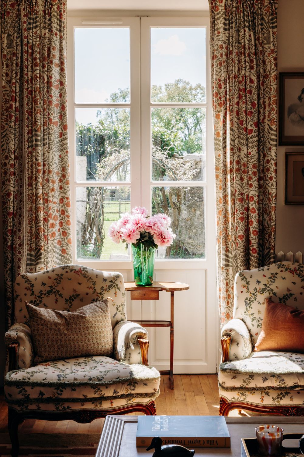 Elegant sitting room with floral armchairs, pink peonies in green vase, and French windows with botanical curtains.