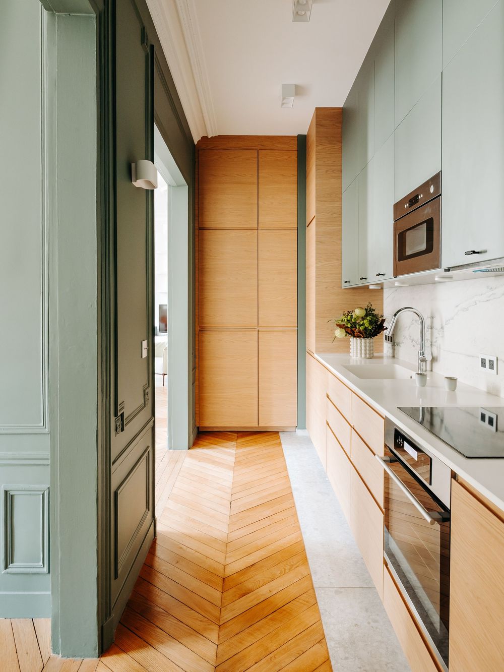 Narrow Parisian kitchen with sage green cabinets, warm wood panels, herringbone floor, marble countertop, and built-in oven.