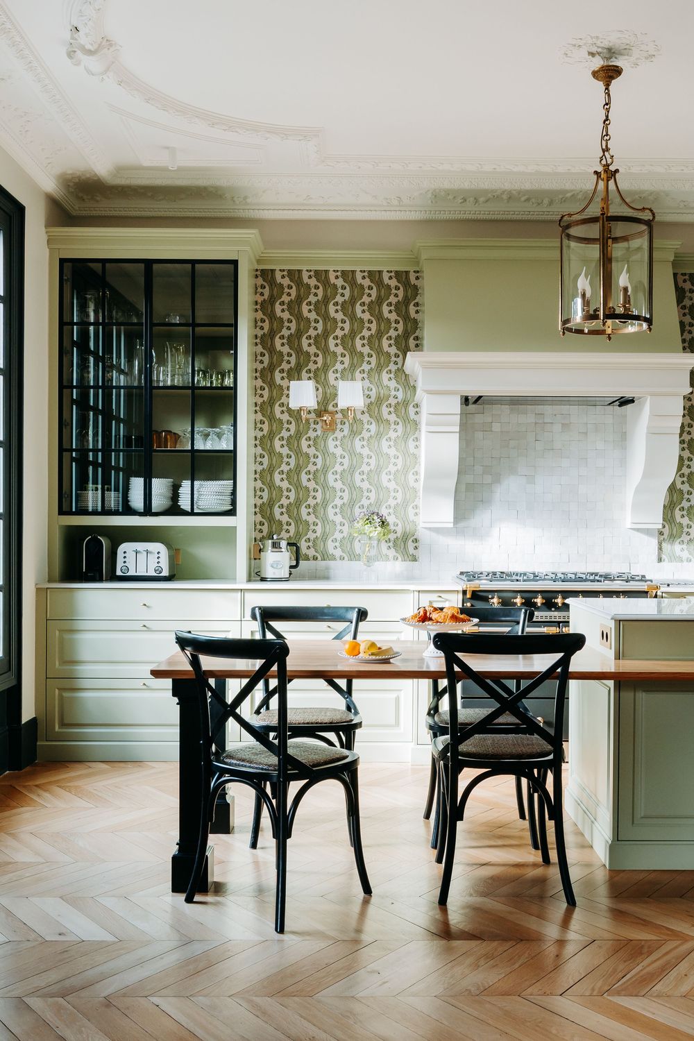 Elegant kitchen with sage green cabinets, herringbone wood floors, brass chandelier, patterned wallpaper, and black bistro chairs at island.
