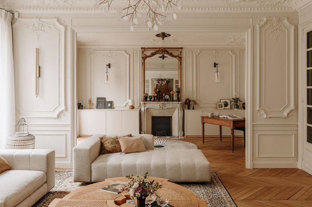 Elegant Parisian living room with white moldings, herringbone floors, neutral sofa, ornate fireplace, and gold mirror.