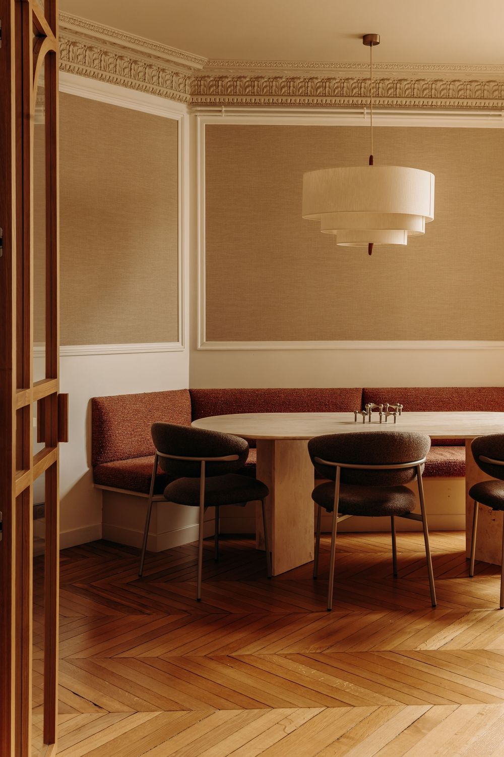 Elegant dining nook with herringbone wood floors, curved banquette, oval table, black chairs, and tiered pendant light.