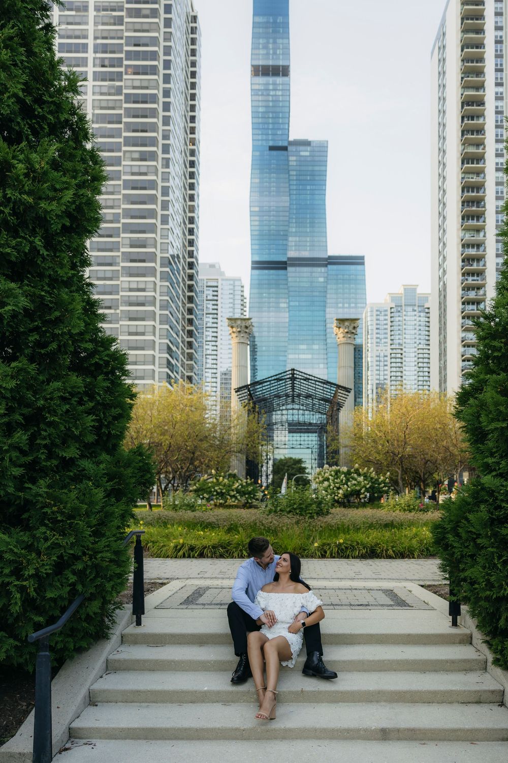 Couple sitting during engagement on steps in Chicago Maggie Daley park with blue glass skyscrapers and lush greenery in the background.