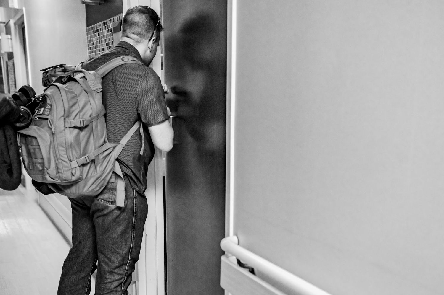 Black and white photo of masked photographer with camera bag entering hospital room door.