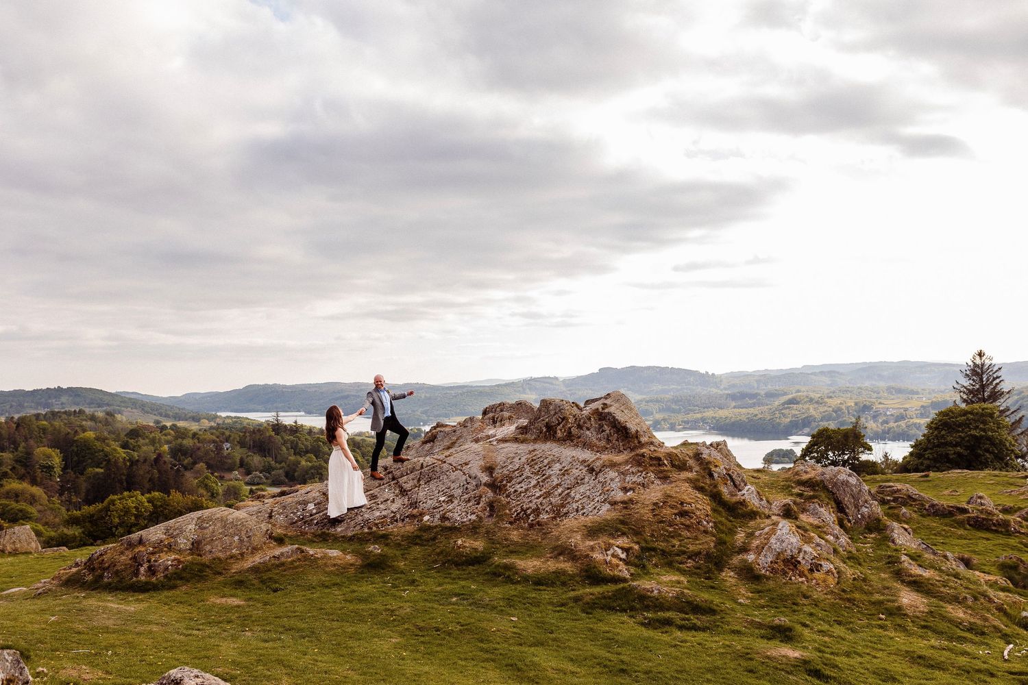 Couple standing on rocky hilltop overlooking a scenic lake and green landscape under a cloudy sky.