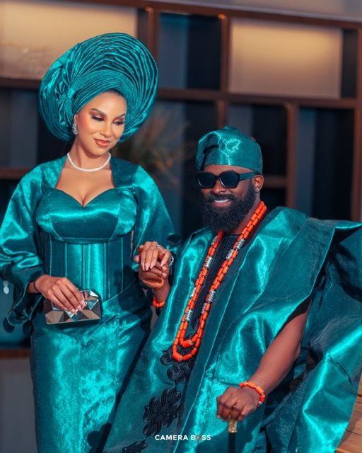 A couple in matching teal traditional African attire with coral beaded necklaces pose together for a formal photograph.