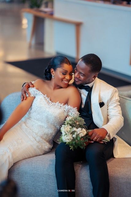 A romantic wedding couple in formal attire share an intimate moment on a gray couch with a white bridal bouquet.