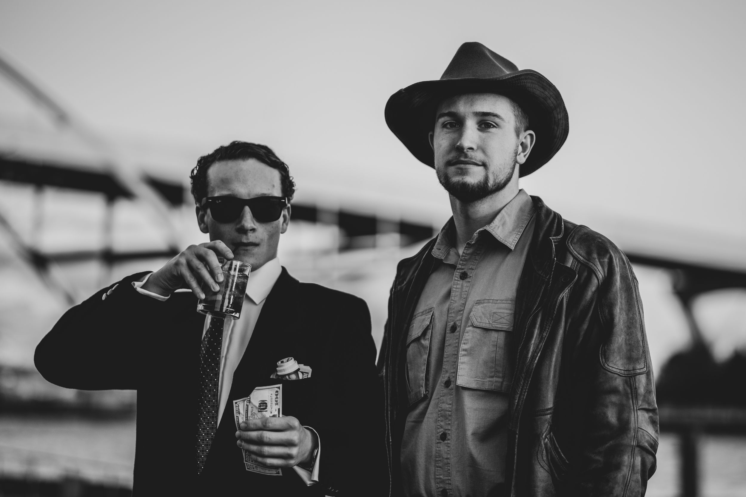 Two men in stylish outfits, one in a suit with sunglasses holding cash, the other in a cowboy hat and leather jacket.