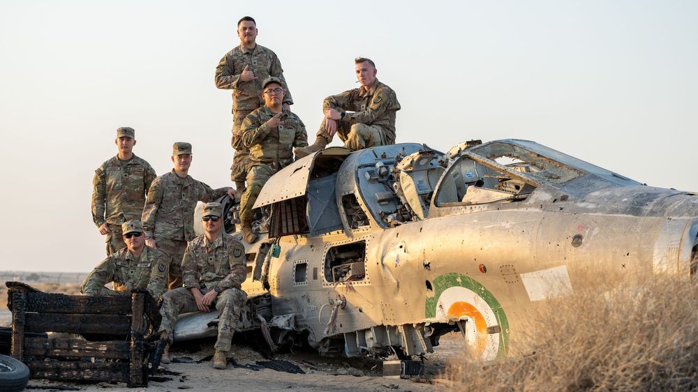 Military soldiers posing on a wrecked fighter jet aircraft in a desert landscape.