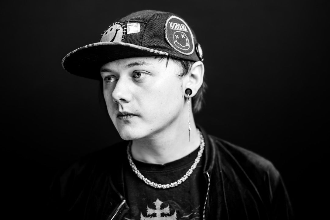 Black and white portrait of a young person wearing a snapback cap, chain necklace, and dark jacket.