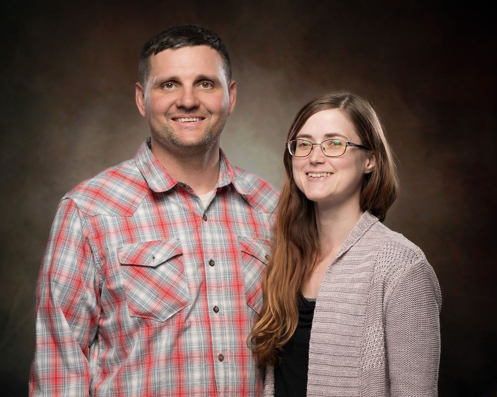A smiling man in a plaid shirt and a young woman with glasses pose together for a professional portrait photo.