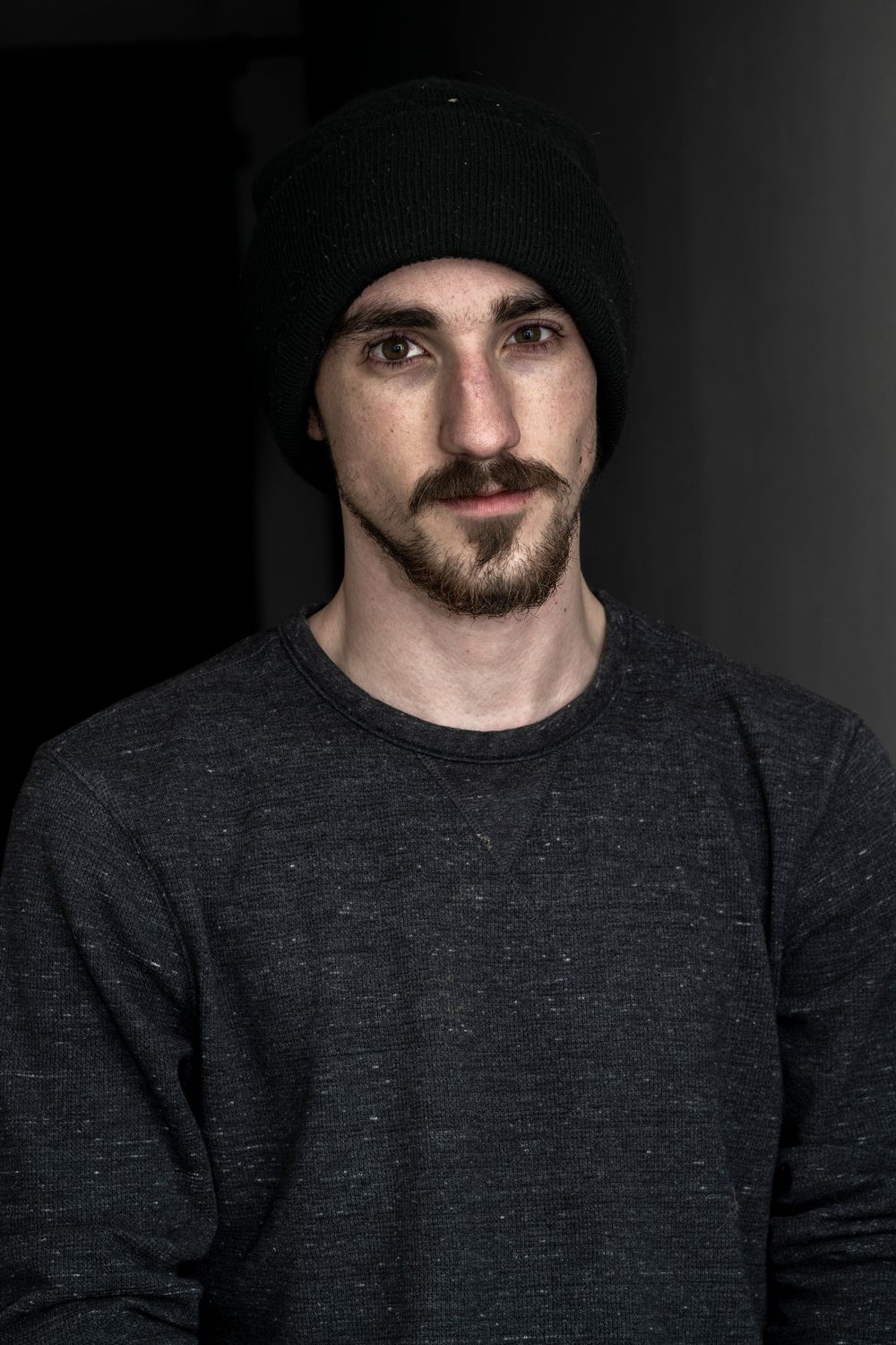 Young man with beard and black beanie hat wearing gray sweatshirt posing against dark background.