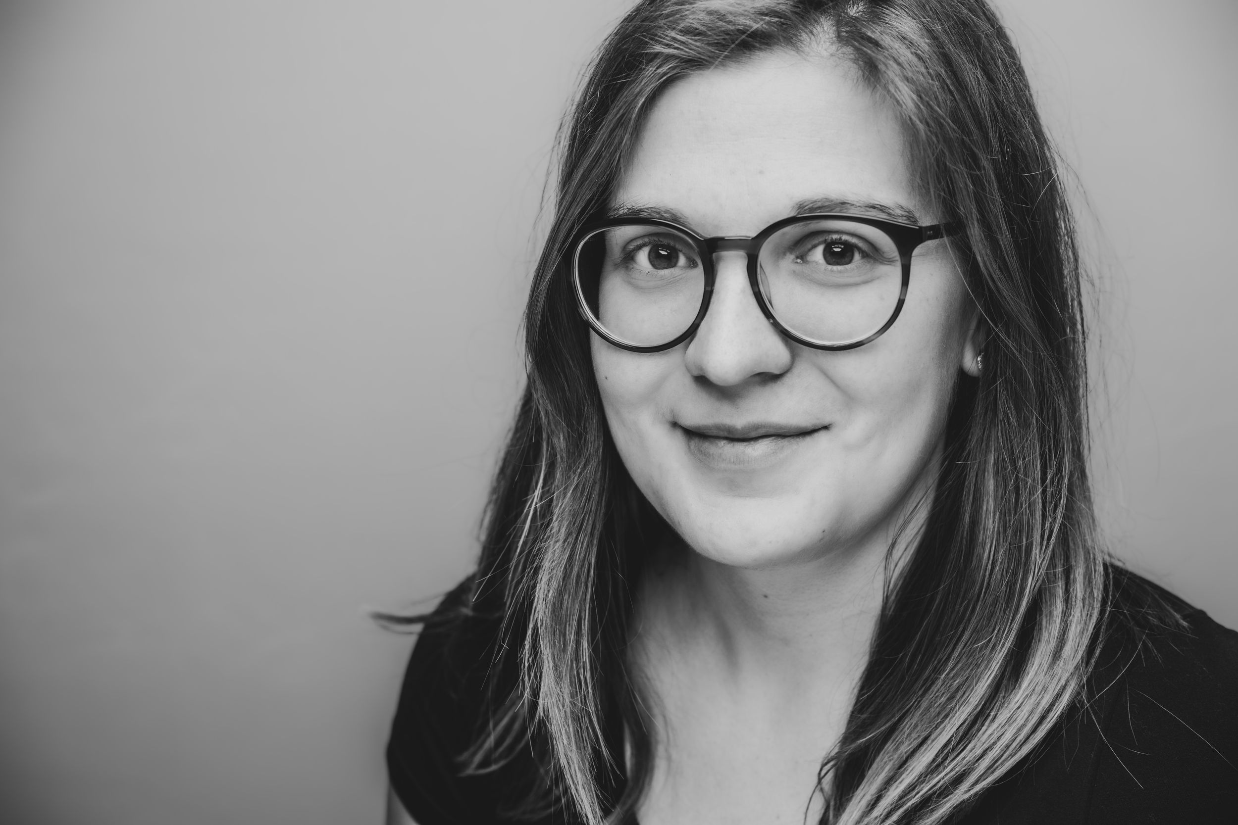 Black and white portrait of a young woman with long hair wearing glasses and a dark top, smiling softly.