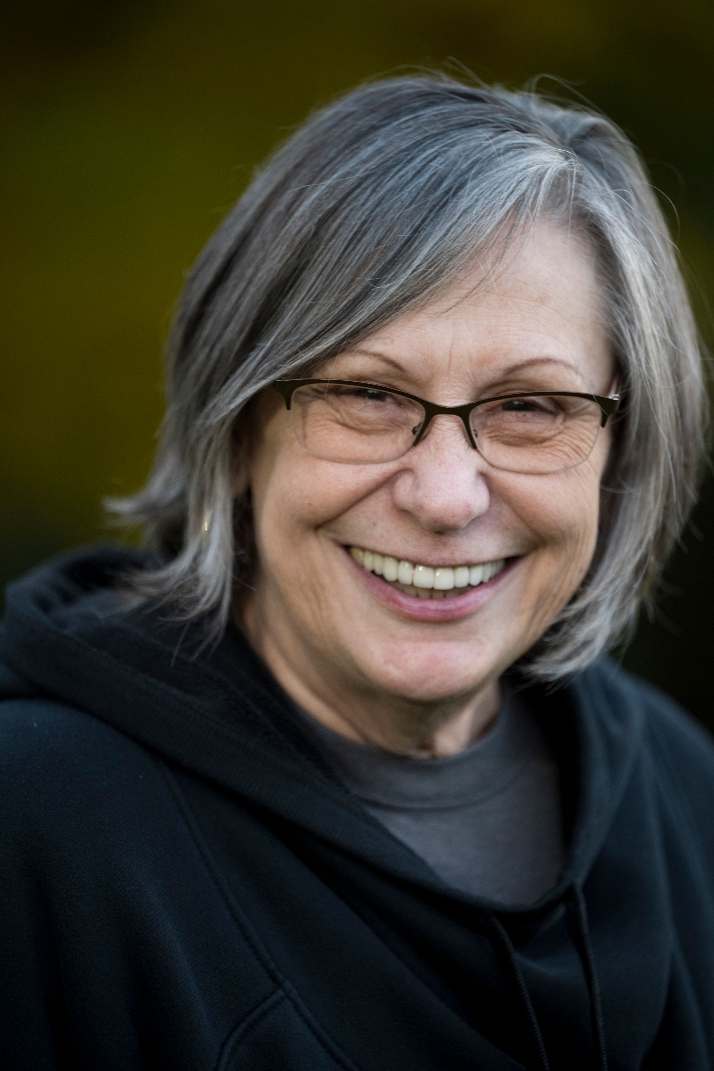 Smiling older woman with gray hair and glasses wearing a black hoodie outdoors.