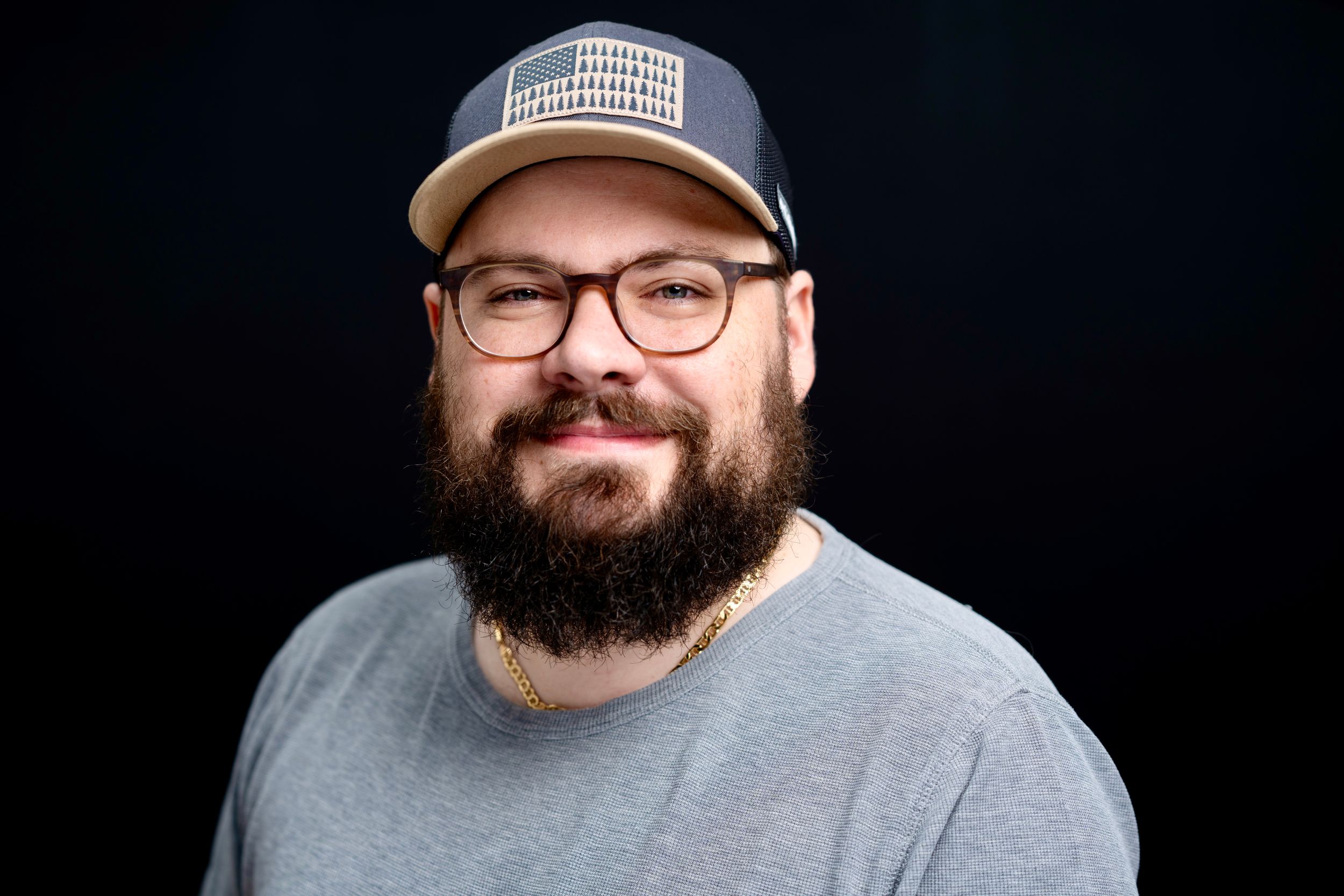 Bearded man with glasses and a trucker hat smiling confidently against a dark studio background.