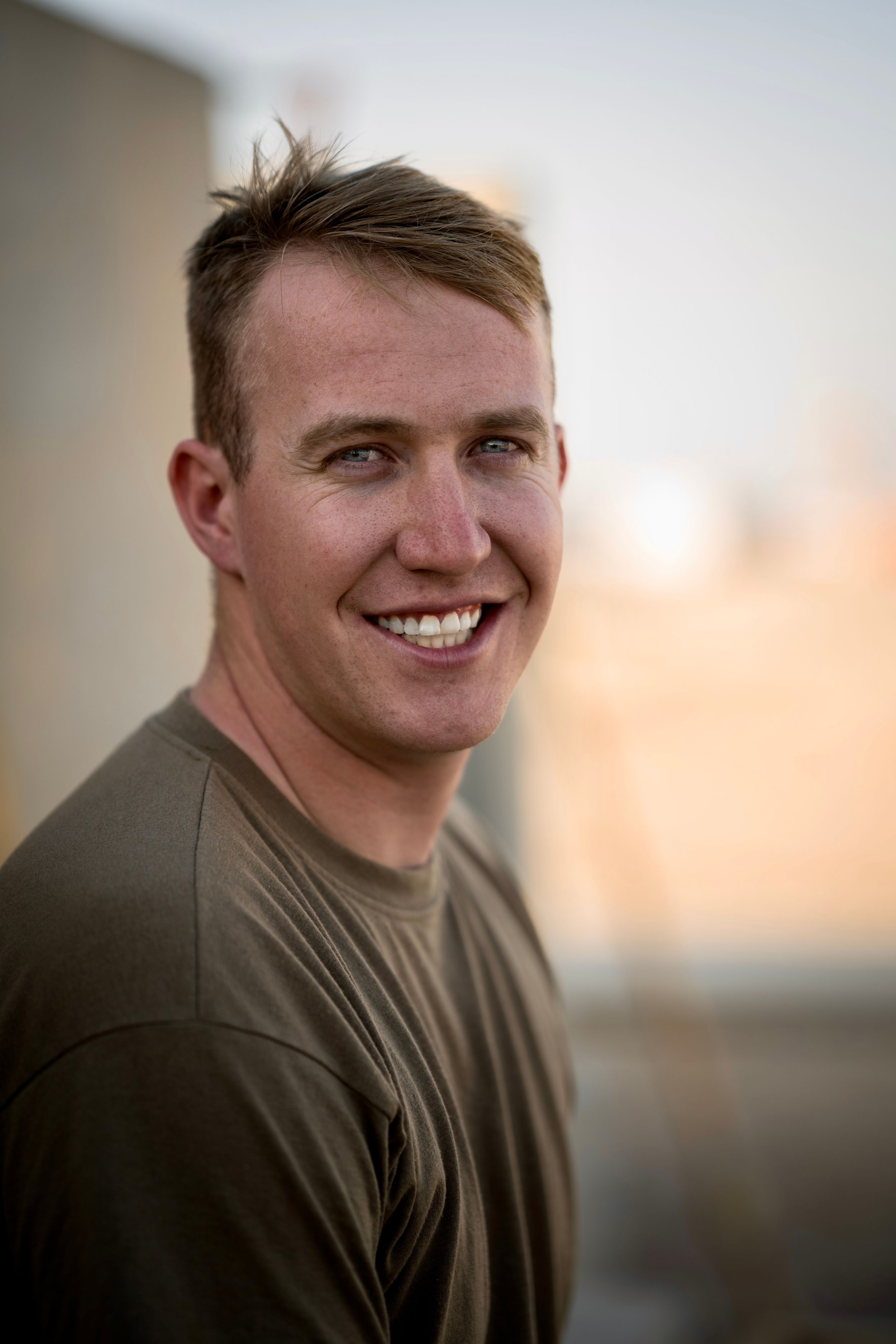 Smiling young man with short hair wearing an olive green t-shirt outdoors with a blurred urban background.