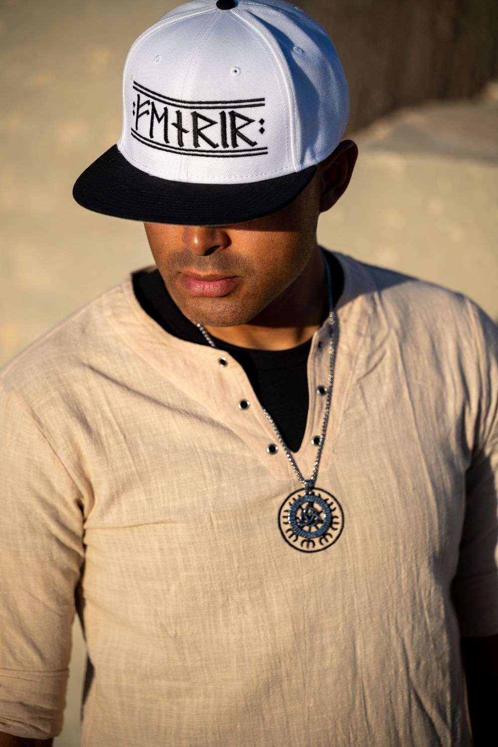 Man wearing white snapback cap and beige linen shirt with circular pendant necklace in warm sunlight.