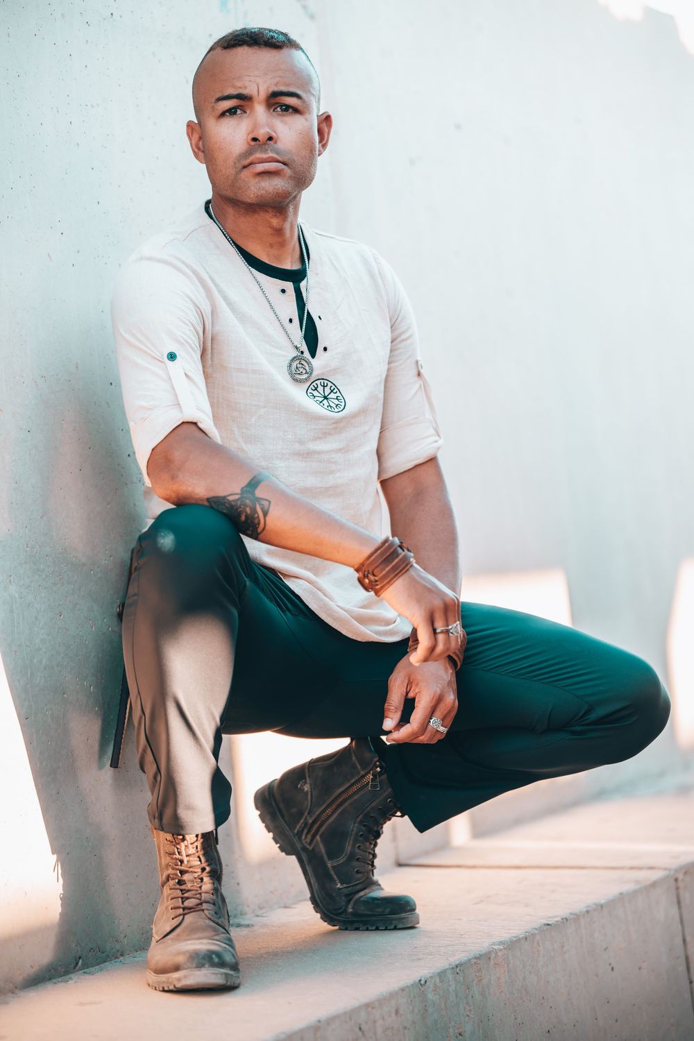 Man in white t-shirt, teal pants, and boots crouching on steps, wearing a pendant necklace and leather bracelets.