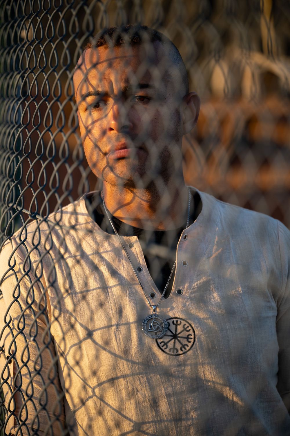 Man in white linen shirt with Viking compass pendant leans against chain-link fence in dramatic golden light.