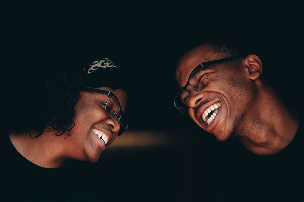 Two people laughing and smiling together in a dark setting, both wearing glasses.