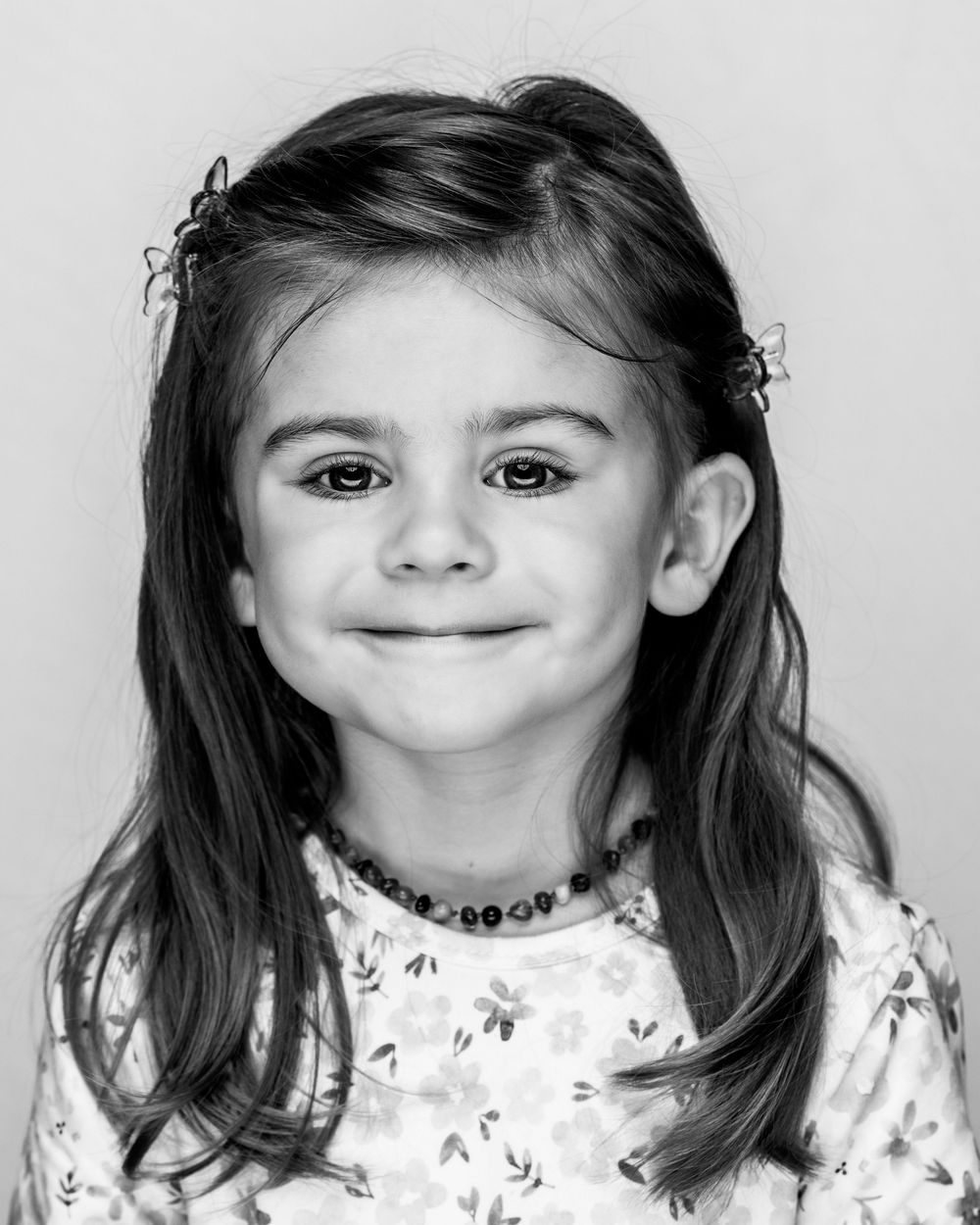 Black and white portrait of a young girl with long hair, hair clips, and a floral shirt, smiling softly.