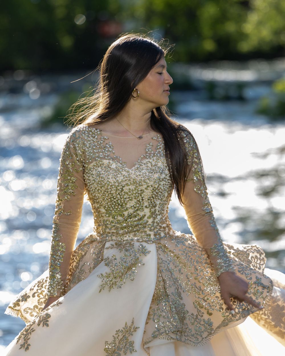 Woman in gold sequined ball gown poses gracefully by a sparkling river in natural sunlight.