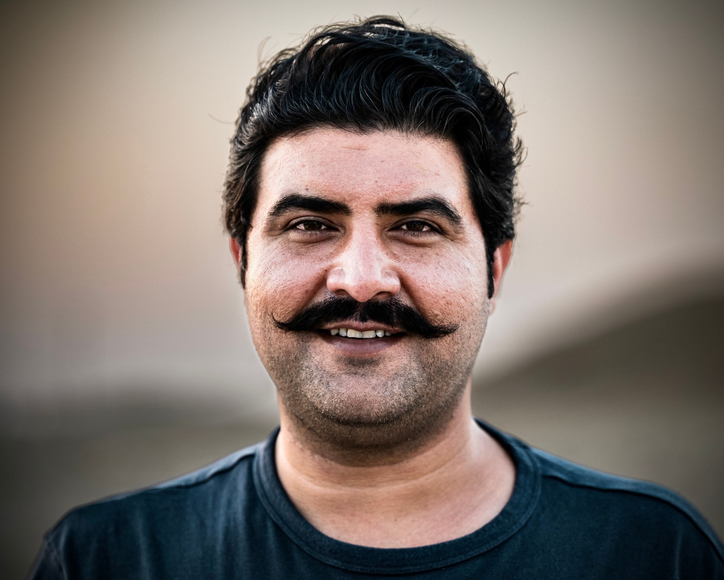 Smiling man with a thick mustache wearing a black t-shirt, portrait photo with blurred background.