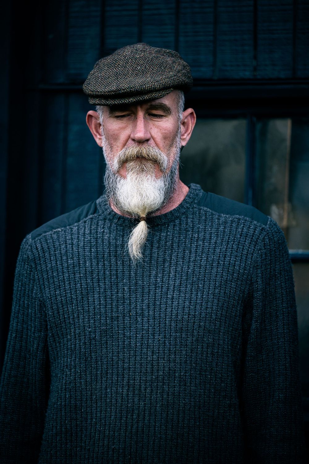 Older man with white braided beard wearing a flat cap and dark knit sweater, looking downward outdoors.