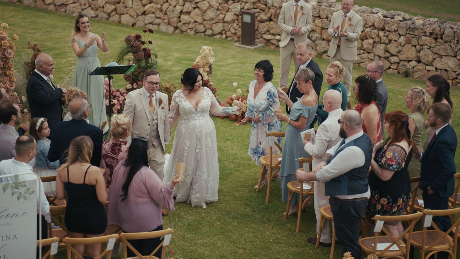 Outdoor wedding ceremony on green lawn with bride and groom exchanging vows surrounded by guests in elegant attire.