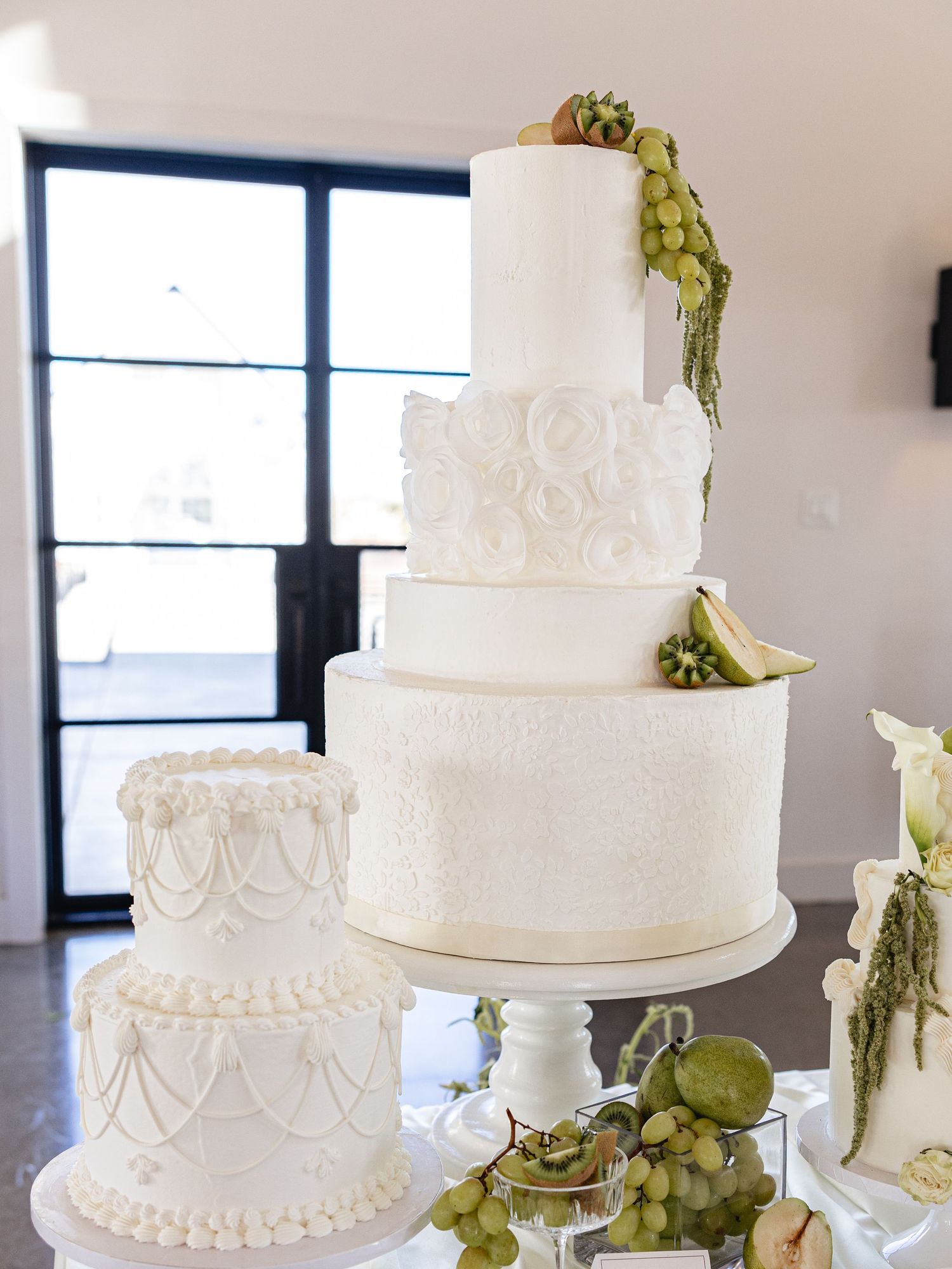 Elegant white multi-tiered wedding cake decorated with roses and fresh green grapes and fruit on a white cake stand.