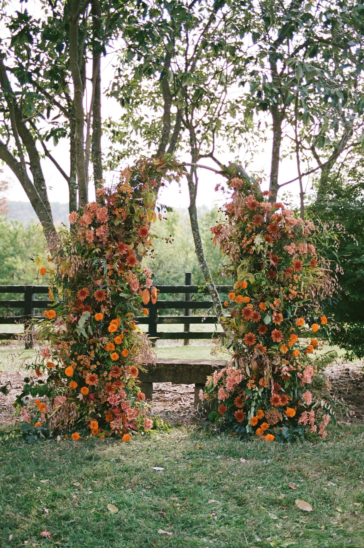 Rustic floral arch with orange and pink blooms flanking a wooden bench outdoors for a fall wedding ceremony.