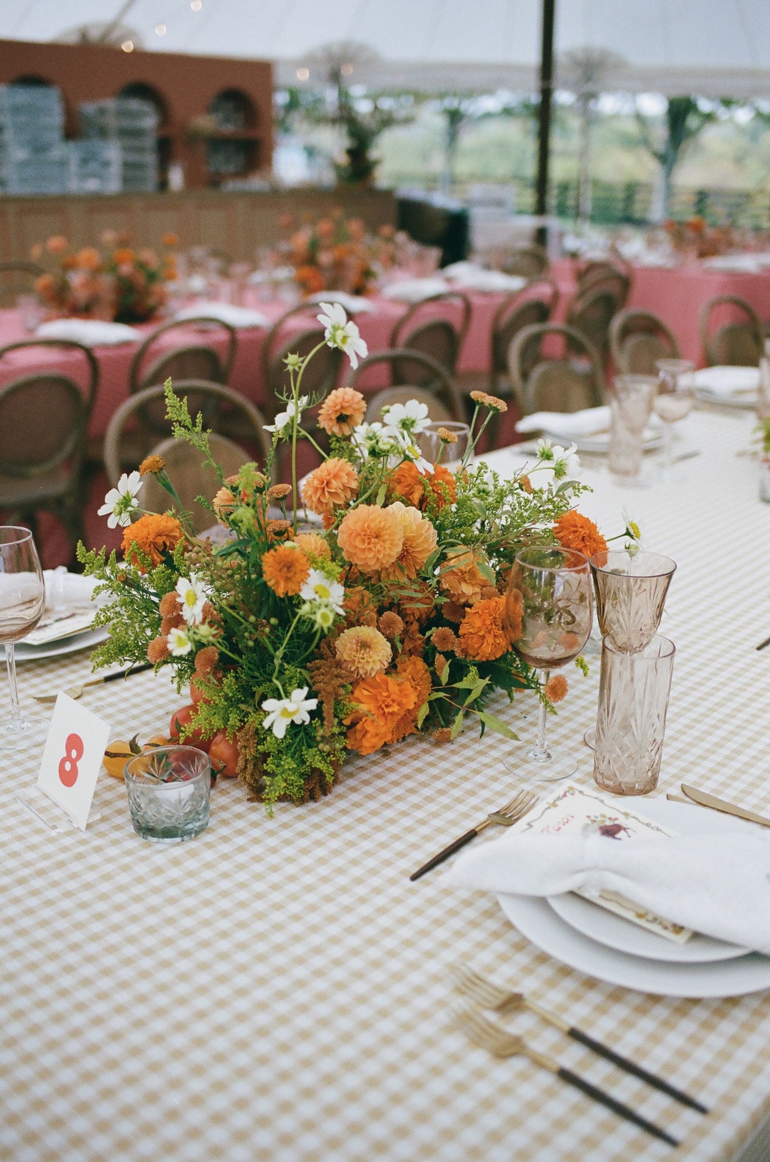 Elegant wedding tablescape with orange marigold centerpiece on gingham linen, gold cutlery, and blush glassware under a tent.