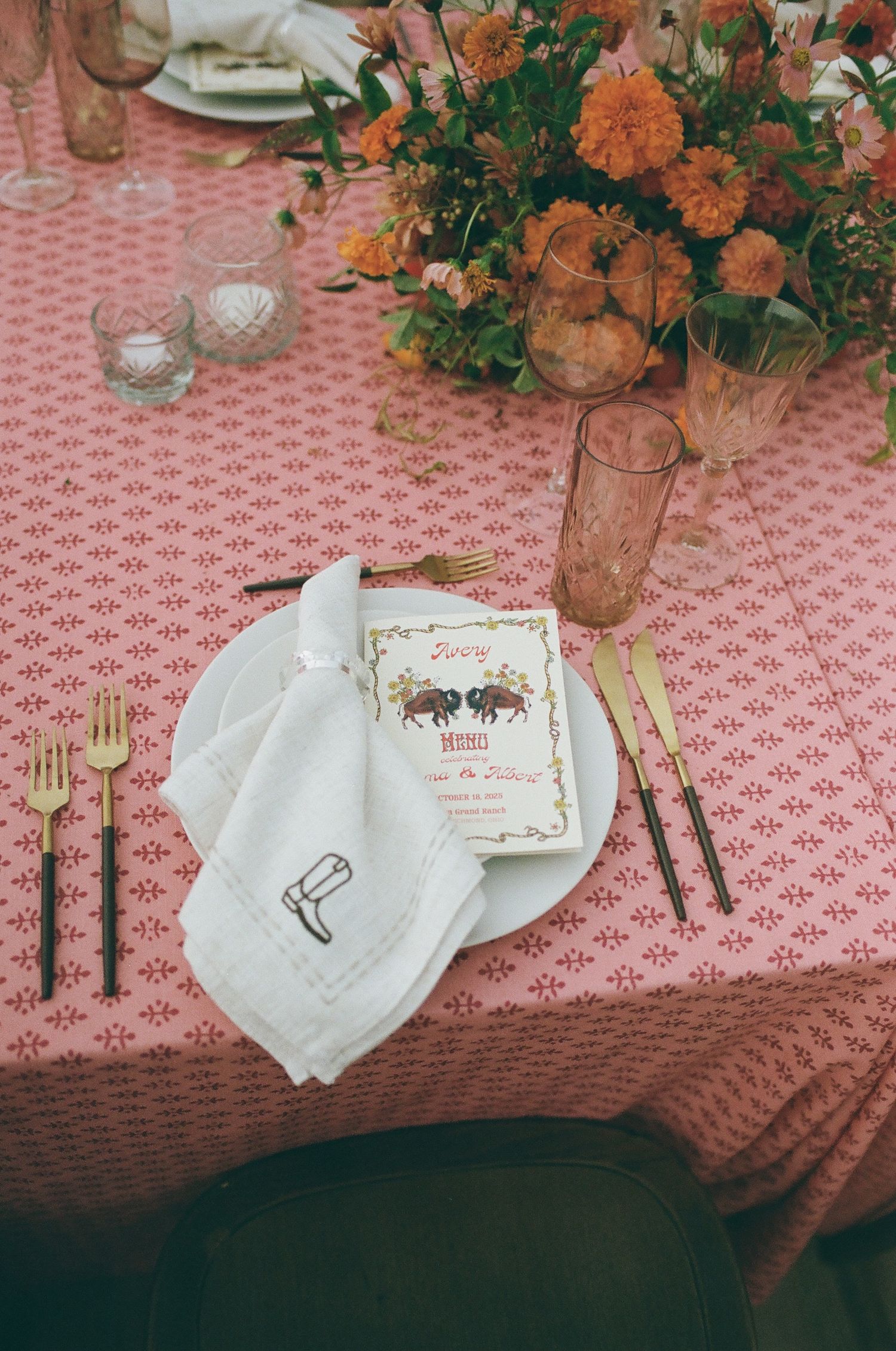 Elegant wedding table setting with pink floral tablecloth, gold cutlery, white napkin, menu card, and orange floral centerpiece.