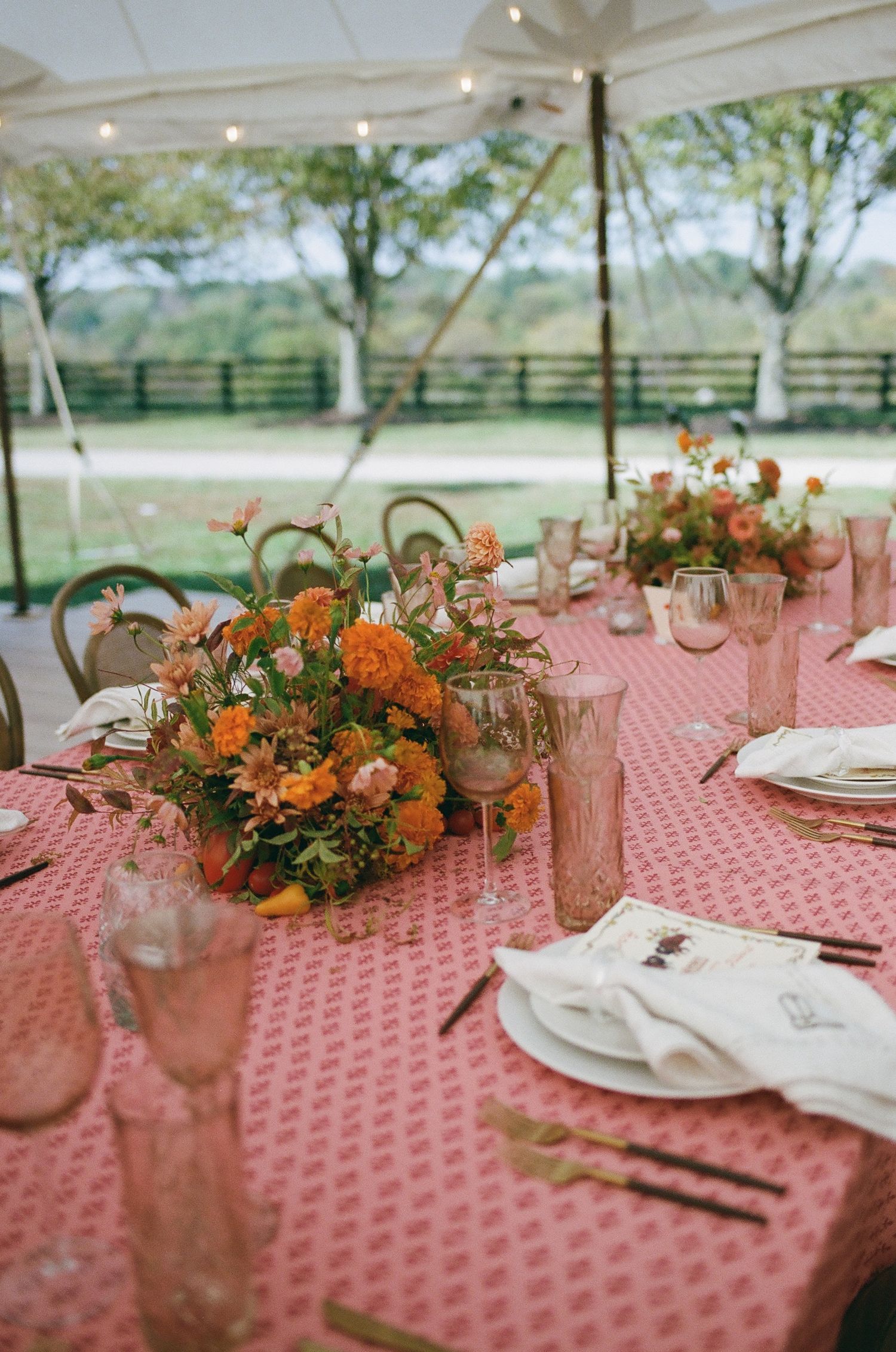 Elegant wedding reception table with pink patterned linen, orange floral centerpiece, and blush glassware under a tent.