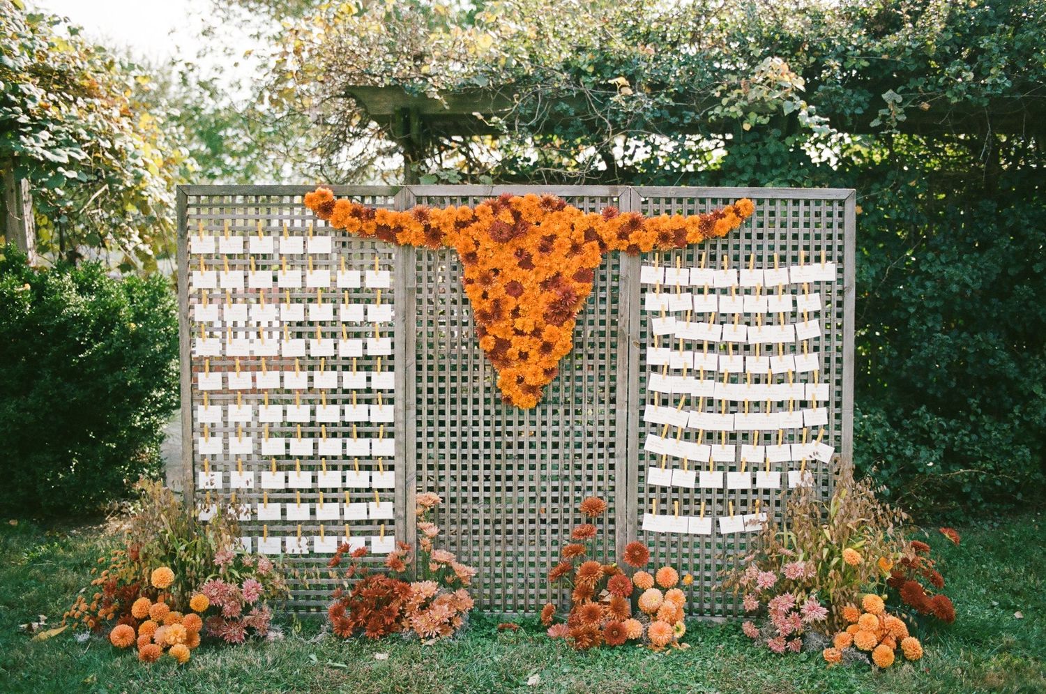 Fall wedding seating chart display with orange floral fabric, lattice panels, and pumpkin floral arrangements outdoors.