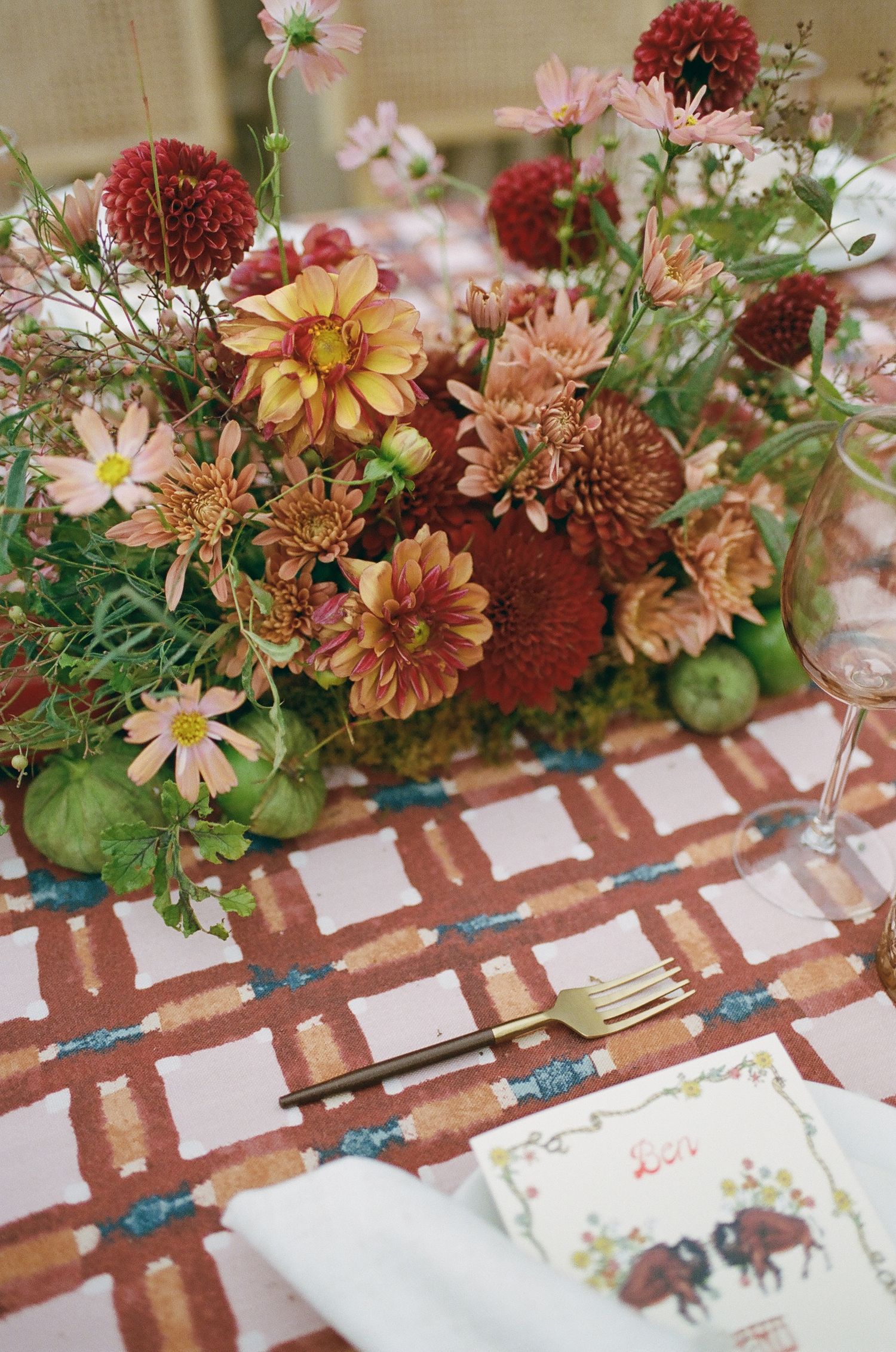 Autumn wedding tablescape with dahlia floral centerpiece, gold fork, patterned rust linen, and place card on elegant table.